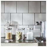 Tre CITRONHAJ krydderiglas i klart glas med metallåg, fyldt med korn, laurbærblade og salt, pænt arrangeret på en bordplade.