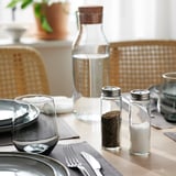 Krydderiglas i klart glas med metalhætter på borddækning, indeholdende kaffegrums og salt, ved siden af en vandkande.