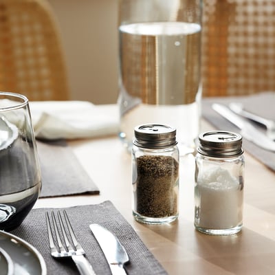 To glaskrukker med metalhætter til sort peber og salt fra ikeas CITRONHAJ serie, på et spisebord med bestik og et vinglas.