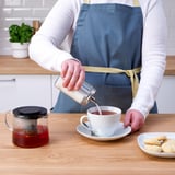 En person, der bærer et forklæde, hælder te i en kop fra en glasbeholder. En tekande med infusionsenhed og småkager står på bordet.