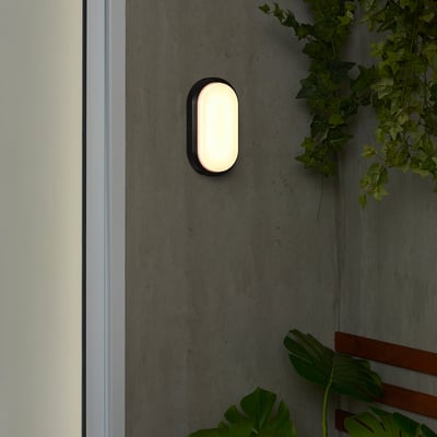 Lille, oval, sort væglampe, DÄCKSBÅT, med polycarbonatskærm, udsender varmt lys, monteret på en grå væg ved siden af grønne planter.