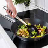 En hånd holder en langskaftet spatel, der rører farverige grøntsager i en non-stick pande på en moderne sort komfur.