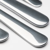 Poleret bestik i rustfrit stål - kniv, ske, gaffel - pænt arrangeret på hvid overflade, der reflekterer lys.