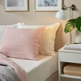 Hyggeligt soveværelse med hvide linnedlagner, pastelpuder, natbord, bøger, lampe, plante og kunst.