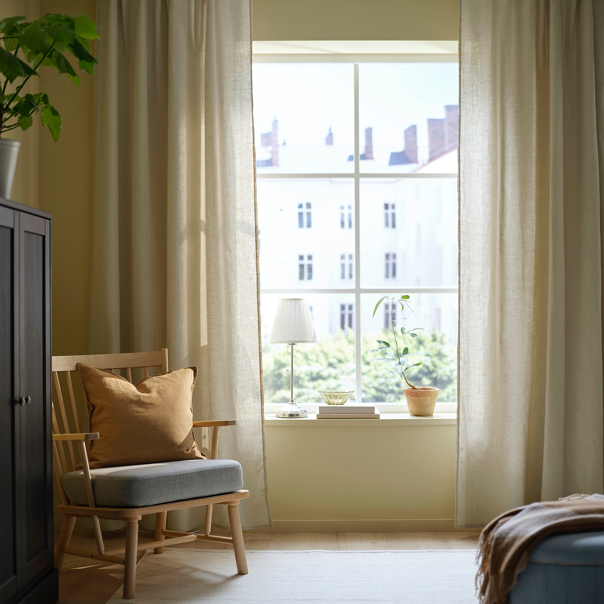 Stort vindue med beige DYTÅG hørgardiner, gyngestol i træ, potteplante, lampe, bøger på karmen.