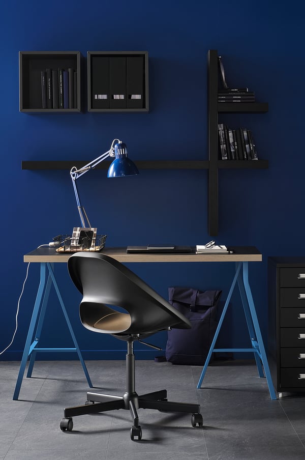 Moderne arbejdsplads med blåt tema med sort ELDBERGET-stol, blå lampe og mørke hylder med bøger og mapper.