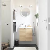 Moderne badeværelse med lyst egetræsskab ENHET, hvid håndvask, rundt spejl og grå håndklæder.