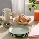 Borddækning med lysegrønne tallerkener, beige skåle, hvide krus, rundstykker, syltetøj, juice og lyserøde servietter.