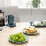 Borddækning med FÄRGKLAR sidetallerkener, kopper og mad. Et hyggeligt måltid med brød, vindruer og kaffe.