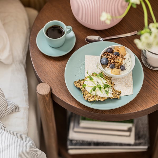 Træbordet har en mintfarvet tallerken med mad og en kaffekop. Der er en seng med puder ved siden af.