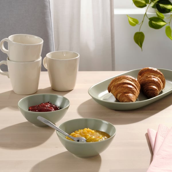 Morgenmadsopsætning med hvide krus, croissanter og marmelade og honning i grønne skåle på træbord.