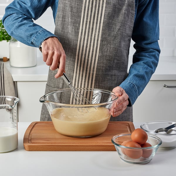 Person blander dej med FINMAT piskeris i glasskål på skærebræt, omgivet af ingredienser.