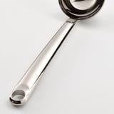 Suppeske i rustfrit stål med buet håndtag og hængende hul, der ligger på en hvid overflade.