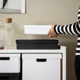Hånden rækker ud efter hvid FJÄDERHARV-kasse oven på skrivebordet, ved siden af sort boks og karaffel, der understreger kassernes størrelse og moderne design.