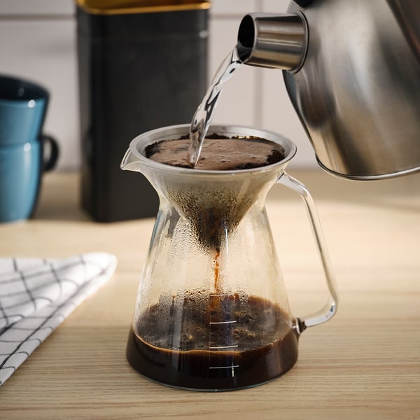 En klar glaskaffedrypper, FLÖJT, hælder varmt vand over kaffegrumsene og skaber en bryg. På en træbordplade.