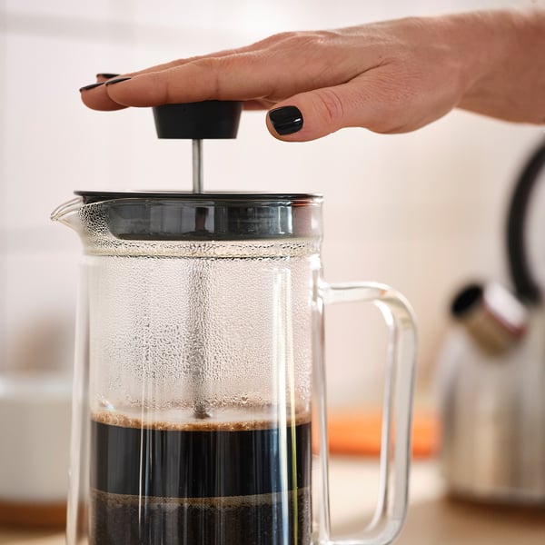 Hånden trykker ned på en cafetière fyldt med kaffegrums, og trækker brygget kaffe ud.