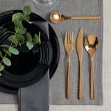 Elegant FÖRNUFT bestiksæt: sorte håndtag, skinnende guld, rustfrit stål, enkelt design. Godt placeret på grå dug, ved siden af sort tallerken og blade.