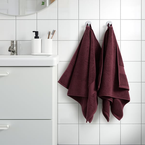Maroon FREDRIKSJÖN-håndklæder hænger på badeværelseskroge, omgivet af minimal indretning med hvide fliser og forfængelighed, der viser blød bomuldstekstur.