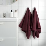 Maroon FREDRIKSJÖN-håndklæder hænger på badeværelseskroge, omgivet af minimal indretning med hvide fliser og forfængelighed, der viser blød bomuldstekstur.