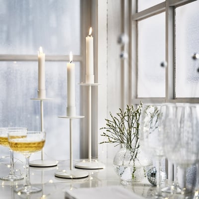 Tre hvide FRÖJDA lysestager på et bord med glas, lys og en vase.