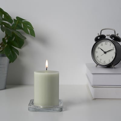 Et tændt stearinlys i en glasholder ved siden af en stak papirer og et ur på en hvid overflade, der tilføjer varme og stemning til omgivelserne.