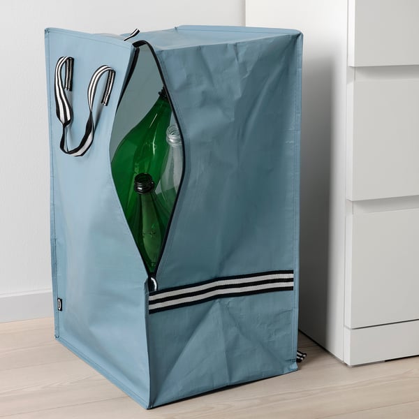 Blå taske med stribede håndtag, lynlås foran åben og viser grøn flaske indeni, læner sig op ad hvidt skab.