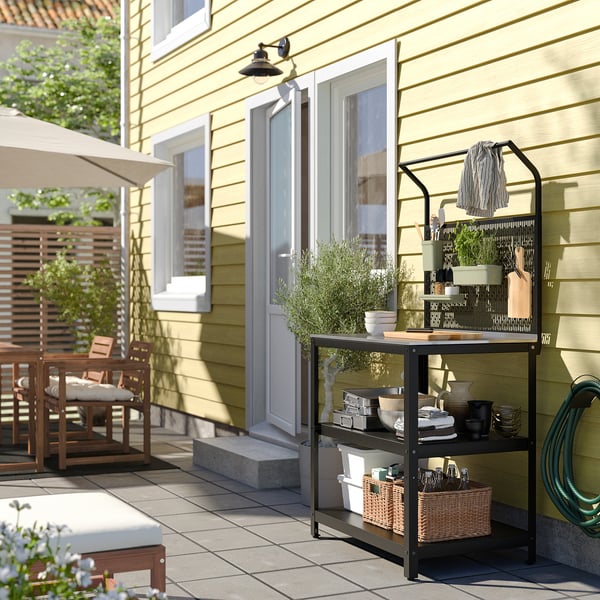Udendørs køkkenopsætning med GRILLSKÄR-vogn med redskaber, tæt på et gult hus og terrassebord.