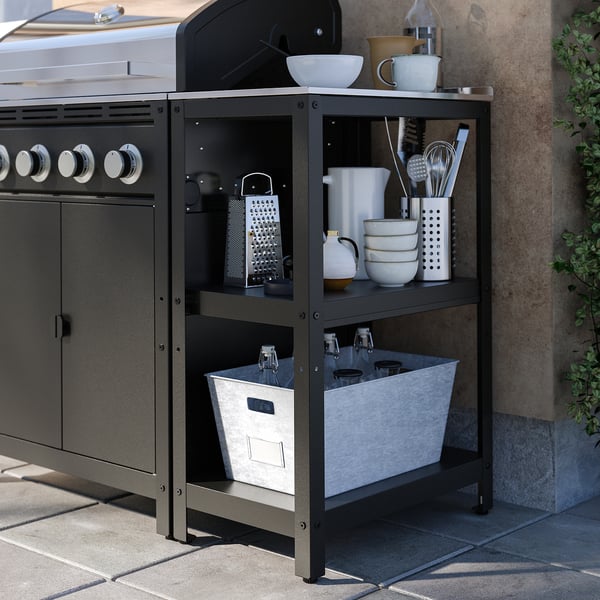 Udekøkken med sort GRILLSKÄR-grill, hylder fyldt med hvid keramik, redskaber og en stofkurv med glasflasker.