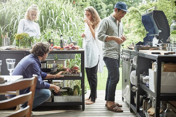 Fire personer laver mad udendørs på en gasgrill. En griller, en hakker grøntsager, to assisterer. Sølvgrill på dæk med grønt.