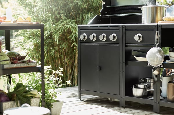 Udekøkken med sort GRILLSKÄR-grill, knopper i rustfrit stål og forskellige køkkenredskaber på hylder, omgivet af potteplanter.