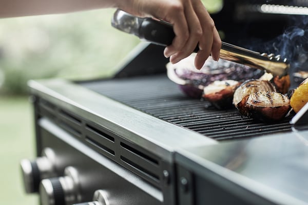 Håndgrillning af mad på GRILLSKÄR sølvfarvet gasgrill i rustfrit stål med sorte betjeningsknapper; forskellige kød synlige.