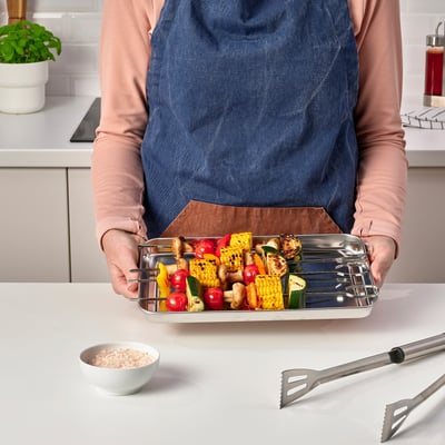 Person, der holder en bakke i rustfrit stål fyldt med farverige grillede grøntsager og spyd.