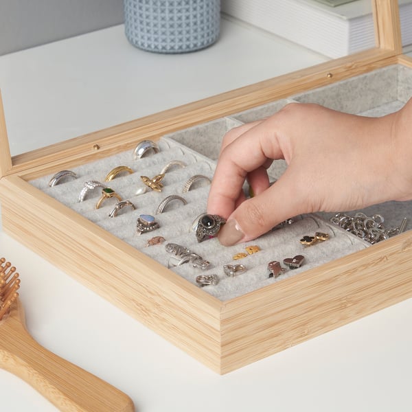 En hånd, der vælger en ring fra et smykkeskrin i træ med rum, der viser forskellige ringe og små accessories.