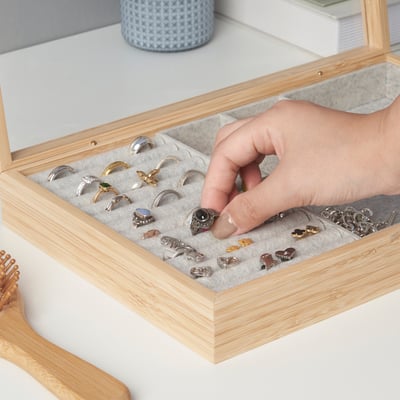 En hånd, der vælger en ring fra et smykkeskrin i træ med rum, der viser forskellige ringe og små accessories.