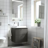 Moderne badeværelse med mørkegrå HAVBÄCK vaskeskab, semi-forsænket vask, spejlbelysning, træskammel med plante og et hængende håndklæde.