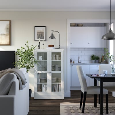 Moderne stue med hvidt fyrretræsskab, glasdøre. L-formet hvidt køkken, grå lampe, sort spisebord med hvide stole.