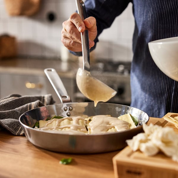 Person koger pasta i en stegepande i rustfrit stål. Sauce hældes over, hvilket fremhæver panderne jævn varmefordeling.