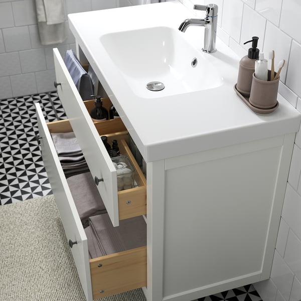 Badeværelsesvask med åbne hvide skuffer med toiletartikler, såsom sæbedispensere og håndklæder, og en vask med en kromarmatur.