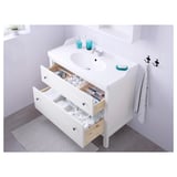 Hvidt badeværelsesbord med åbne skuffer med organiserede toiletartikler og håndklæder. Vask med blå beholdere og krog med håndklæder ved siden af.
