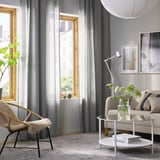 Moderne stue med store grå HILJA gardiner, beige sofa, rundt sofabord og træstol ved vinduer.