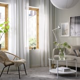 Moderne stue med hvide HILJA gardiner, træstol, grå sofa, rundt sofabord og gråt tæppe.