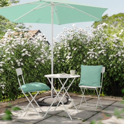 Lille udendørs bordsæt med lysegrønne polstrede stole og mintparasol, omgivet af blomstrende buske.