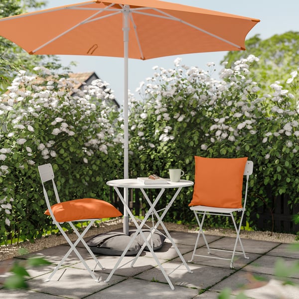 Udendørs terrassesæt med hvidt metalbord og stole, orange hynder og paraply, omgivet af blomstrende buske.