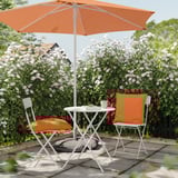 Udendørs terrassesæt med hvidt metalbord og stole, orange hynder og paraply, omgivet af blomstrende buske.
