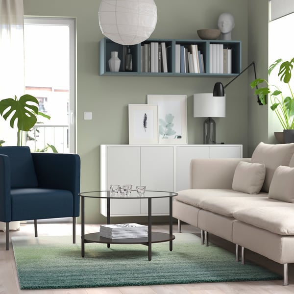 Moderne stue med beige sofa, blå stol, glasbord, grønt tæppe, bogfyldt hylde, grøn accentvæg, kunstværk.