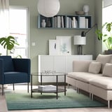 Moderne stue med beige sofa, blå stol, glasbord, grønt tæppe, bogfyldt hylde, grøn accentvæg, kunstværk.