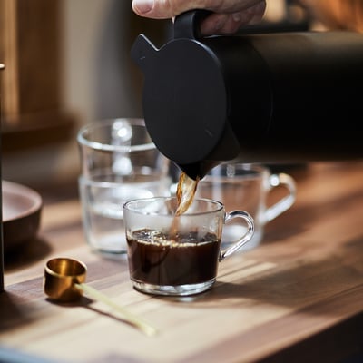 Person hælder kaffe i klart glaskrus på træbord. Krusdel af IKEA 365+ serien, designet til holdbarhed og alsidighed.