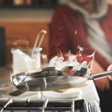En person laver mad i en stegepande i rustfrit stål på en komfur, udsender damp og viser dens jævne varmefordeling og non-stick egenskaber.