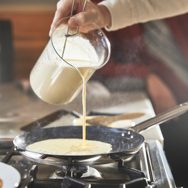 En person hælder dej fra en hvid kande i en non-stick stegepande på et gaskomfur, hvilket illustrerer pandernes jævne varmefordeling og brugervenlighed.