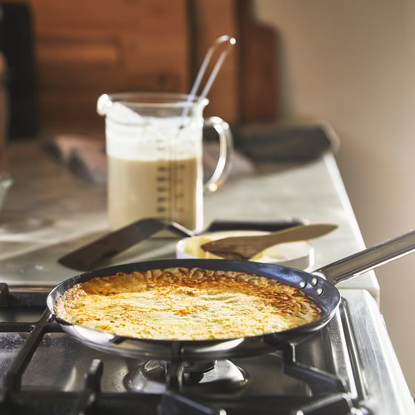 Madlavning: et gaskomfur med en non-stick pande indeholdende en gyldenbrun pandekage. Bagved er der et målebæger med dej og spatel på bordpladen.